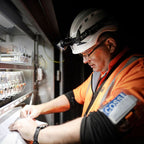 A man in a white hard hat with a Ledlenser H15R headlamp, wearing an orange safety vest over a black shirt, writing on a clipboard inside an industrial electrical panel room. A blue and white 'COSSI' patch is visible on his sleeve.