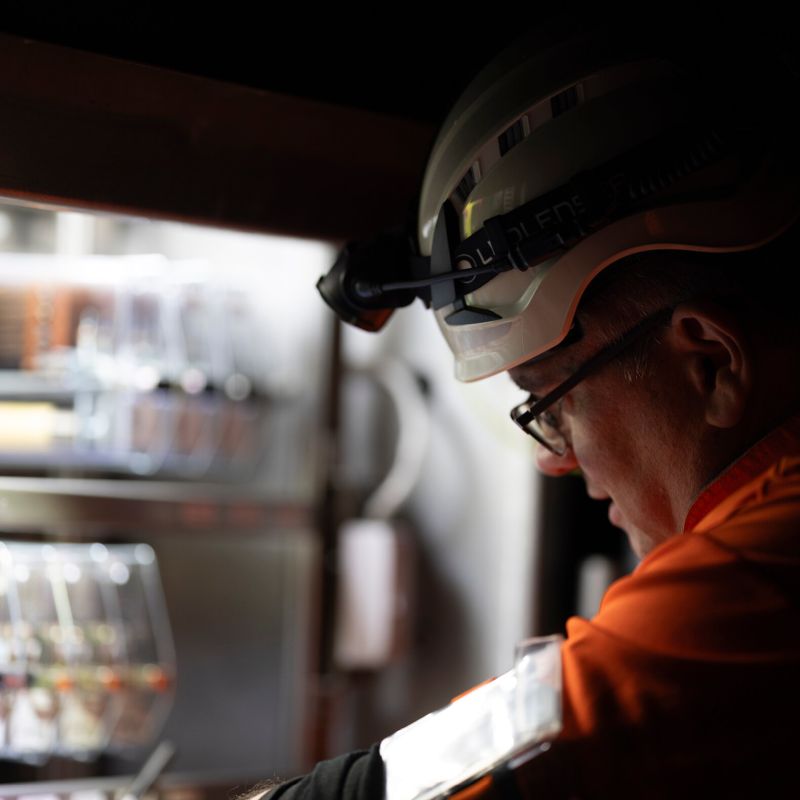 A close-up side profile of a person wearing a white hard hat with a Ledlenser H15R headlamp and a bright orange high-visibility safety jacket, set against a dark, industrial background.