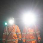 Two workers in high-visibility orange safety jackets with reflective stripes, standing in a dark environment with Ledlenser H14R Pro headlamps turned on, creating strong glare.