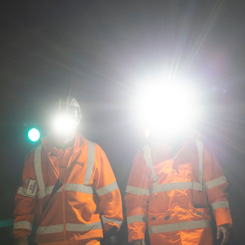 Two workers in high-visibility orange safety jackets with reflective stripes, standing in a dark environment with Ledlenser H14R Pro headlamps turned on, creating strong glare.