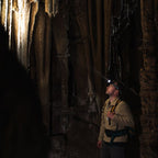 A man wearing a Ledlenser H21 headlamp and an outdoor expedition backpack stands in a dark cave looking up at stalactite formations illuminated by cave lights. 