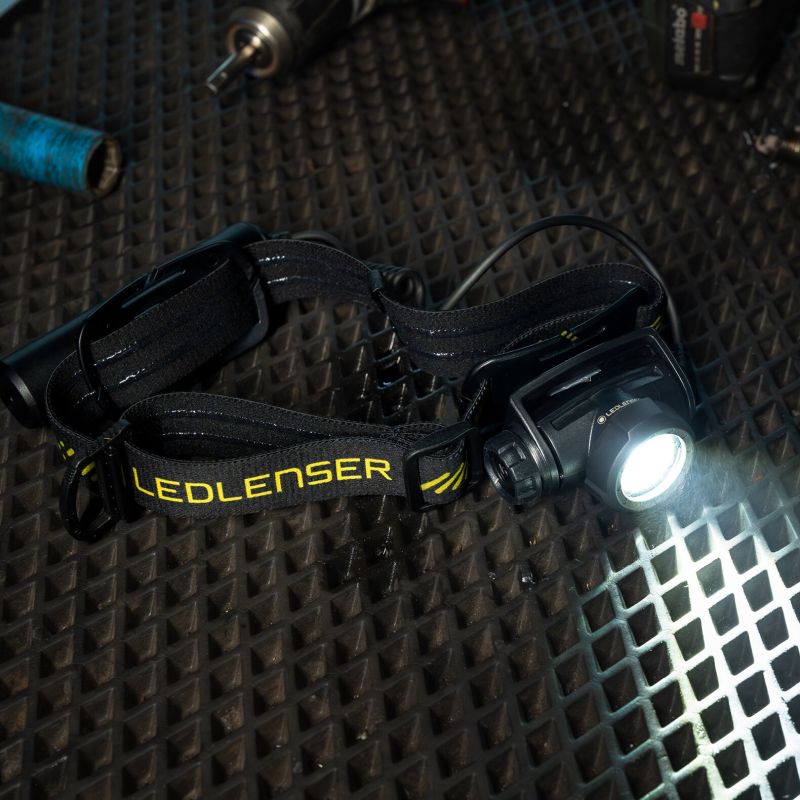 A black Ledlenser H5R Pro headlamp with a bright illuminated light, resting on a dark textured metal surface. The headband features the Ledlenser logo in yellow text.
