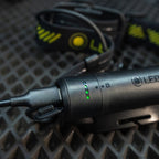 A close-up of a black Ledlenser H5R Pro headlamp showing its charging port and indicator lights, with its matching black and neon yellow strap blurred in the background on a dark textured surface.