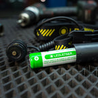 A Ledlenser battery pack with a green and white label, shown in a close-up shot on a textured black surface with an H5R Pro headlamp and other tools blurred in the background.