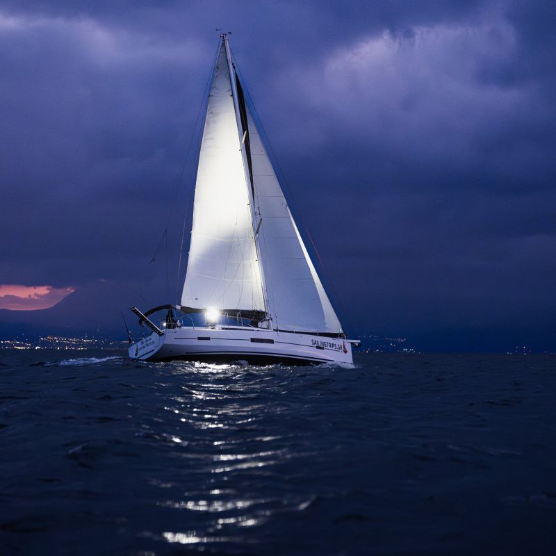 A white sailboat with illuminated sails sailing on dark blue water under a dramatic cloudy night sky.