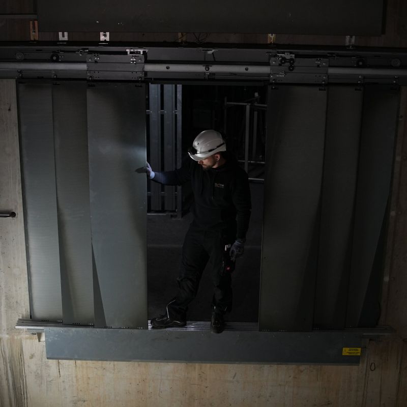 A worker in a white hard hat and dark clothing stands in front of large gray metal sliding doors in a dim industrial setting and using a Ledlenser H149R Pro headlamp to light his way