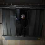 A worker in a white hard hat and dark clothing stands in front of large gray metal sliding doors in a dim industrial setting and using a Ledlenser H149R Pro headlamp to light his way