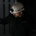 A man wearing a white hard hat with a Ledlenser H19R Pro headlamp and a black hoodie, illuminated in a dark industrial setting.