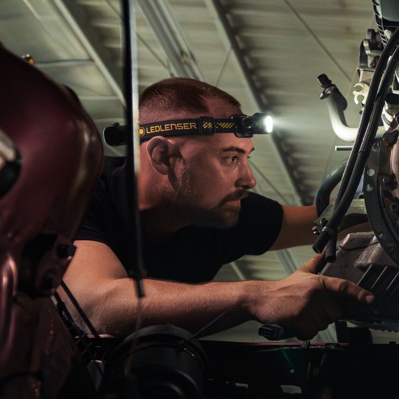 A man wearing a black Ledlenser H7R Pro headlamp with a bright flashlight on, working in a dark industrial or mechanical environment.