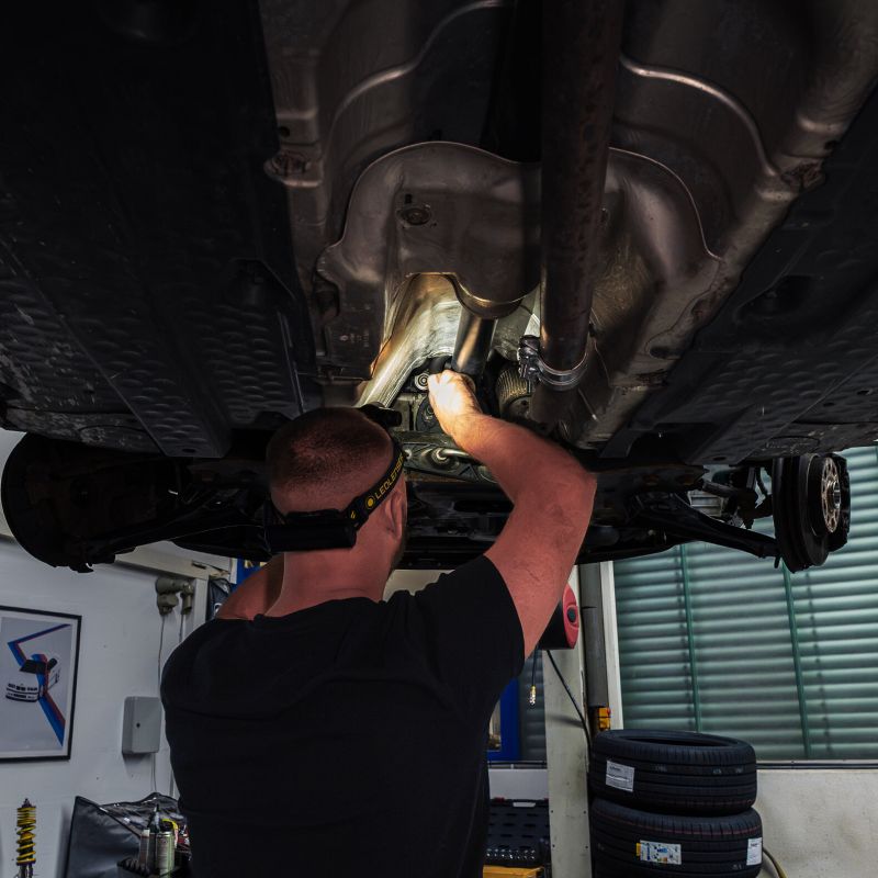 A mechanic wearing a black t-shirt and a Ledlenser H7R Pro headlamp works underneath a car in a garage, illuminating the vehicle's undercarriage with a flashlight.