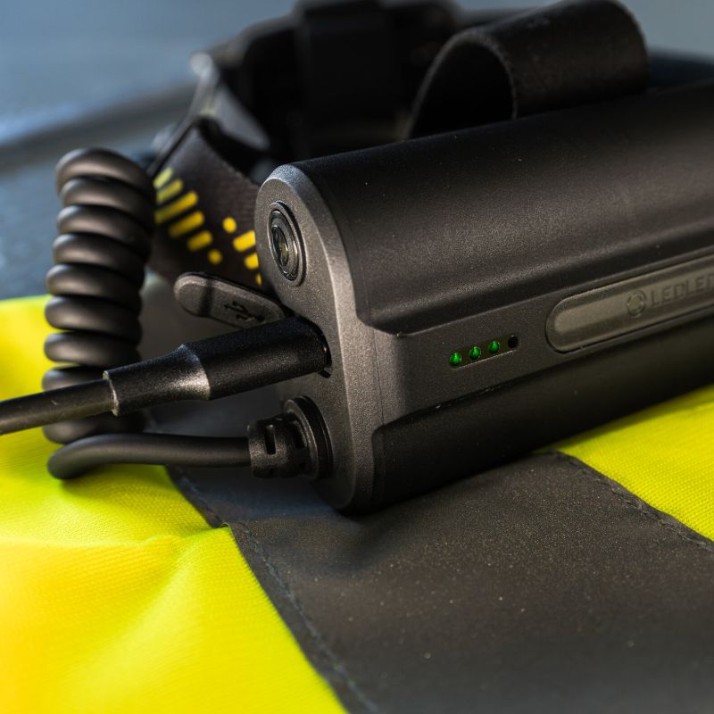 A close-up of a black Ledlenser H149R Pro headlamp battery with a coiled cable, resting on a high-visibility yellow and black safety vest.