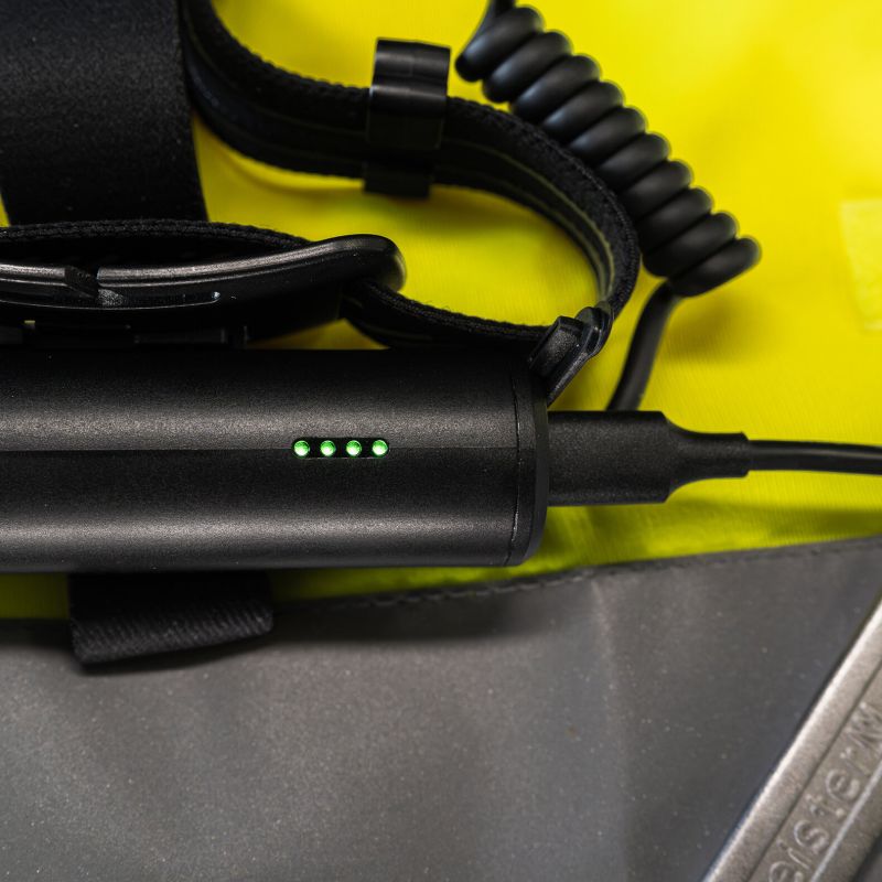 Close-up of a black Ledlenser H7R Pro headlamp with four green LED indicator lights, attached to a coiled black cable, resting on a bright yellow surface.