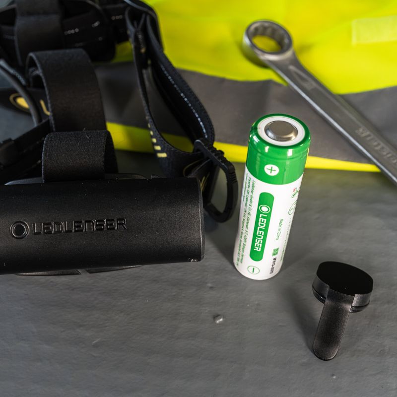 A black Ledlenser H7R Pro headlamp with  battery and its cap next to a black Lumbee headlamp, set against a background of a yellow safety vest and a wrench.
