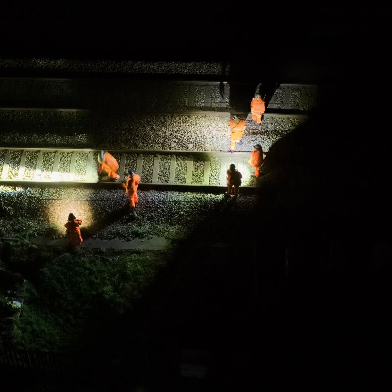 Several workers lighting their way with Ledlenser H19R Pro headlamps in high-visibility orange safety suits standing on railroad tracks at night under harsh overhead lighting.