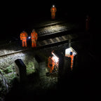 Workers in high-visibility orange safety suits and wearing Ledlenser H15R headlamps, inspecting railway tracks and electrical equipment at night under harsh overhead lighting.