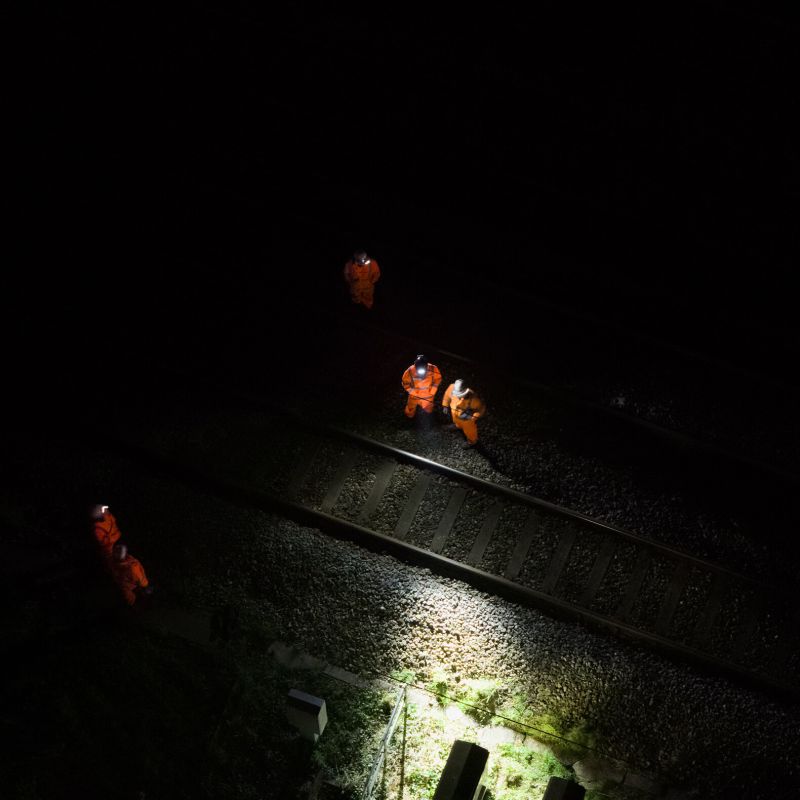 Three workers in high-visibility orange safety suits standing on train tracks at night under harsh overhead lighting, with one worker standing nearby on the left. All are wearing Ledlenser H14R Pro headlamps