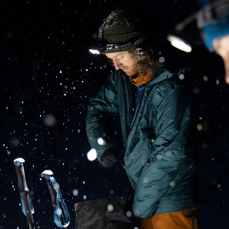 Person in a teal jacket and black beanie standing in a snowy night setting with ski poles nearby and lighting their way with a Ledlenser H19R Signature headlamp