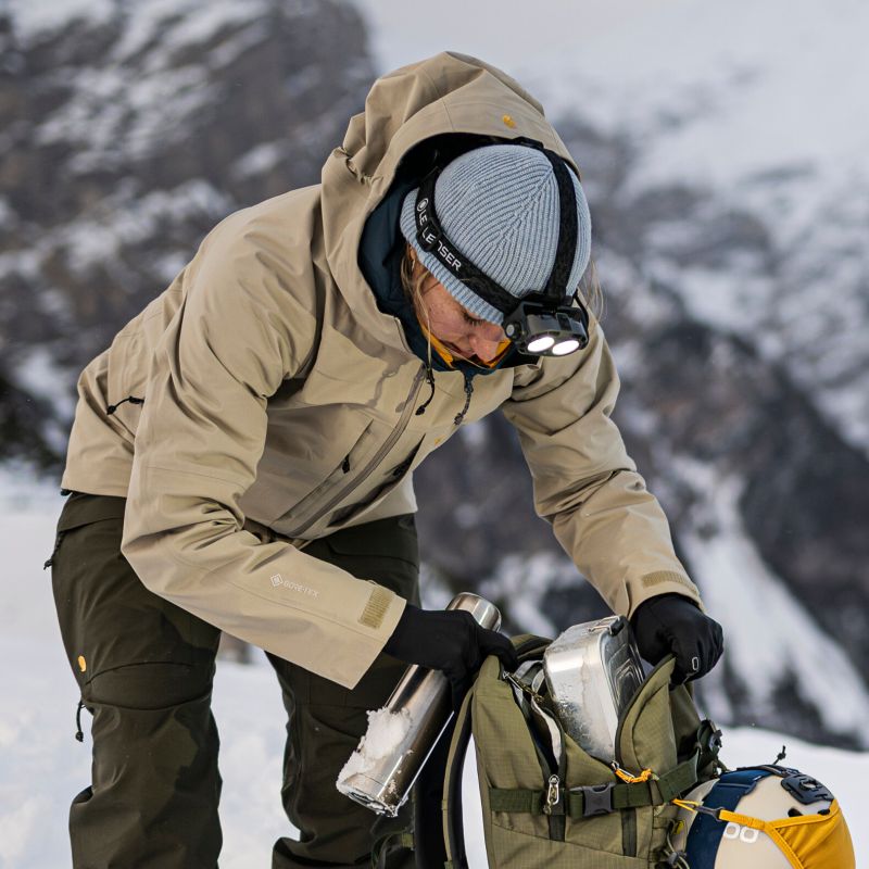 A person in a beige hooded winter jacket, dark pants, a gray knit beanie, and a Ledlenser H21R headlamp packs gear into an olive green backpack in a snowy mountain setting.
