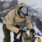 A person in a beige hooded winter jacket, dark pants, a gray knit beanie, and a Ledlenser H21R headlamp packs gear into an olive green backpack in a snowy mountain setting.
