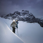 Two winter sports tourists wearing Ledlenser H21R headlamps and ski poles traverse a snowy mountain slope, with a rugged, snow-dusted peak under a dark, moody sky in the background.