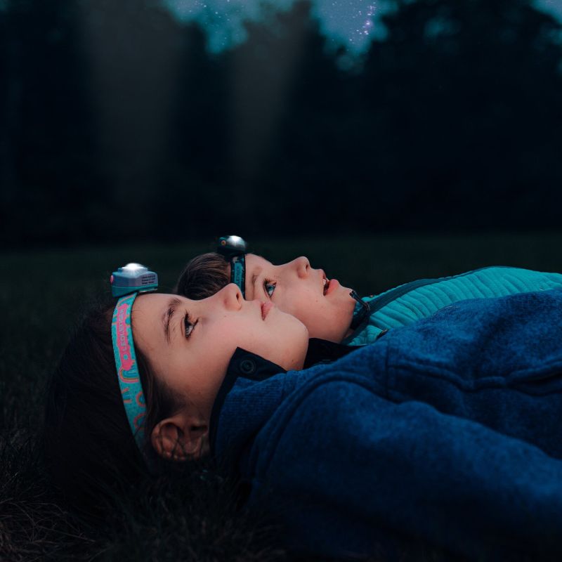 Two young girls laying on their backs wearing Ledlenser KIDLED4R headlamps while out in nature - Rainbows pattern