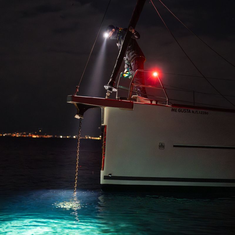 A person on a sailboat at night uses a Ledlenser H19R headlamp to project light into the water, illuminating glowing blue bioluminescent water. The boat's side is visible, with a white hull, and red navigation lights