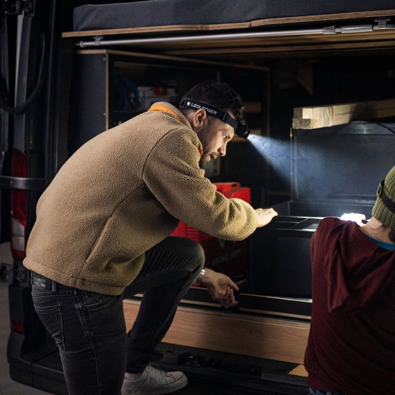 A man wearing a tan fleece jacket and dark jeans crouches inside the open rear cargo area of a vehicle, using a Ledlenser H5R headlamp to illuminate the interior. Another person in a burgundy shirt is partially visible on the right.