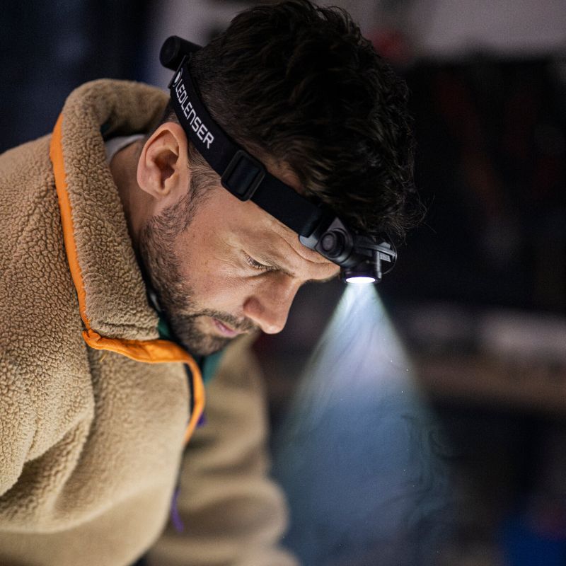 A man wearing a black Ledlenser H5R headlamp with a bright beam of light shining downward, dressed in a brown fleece jacket with orange trim.